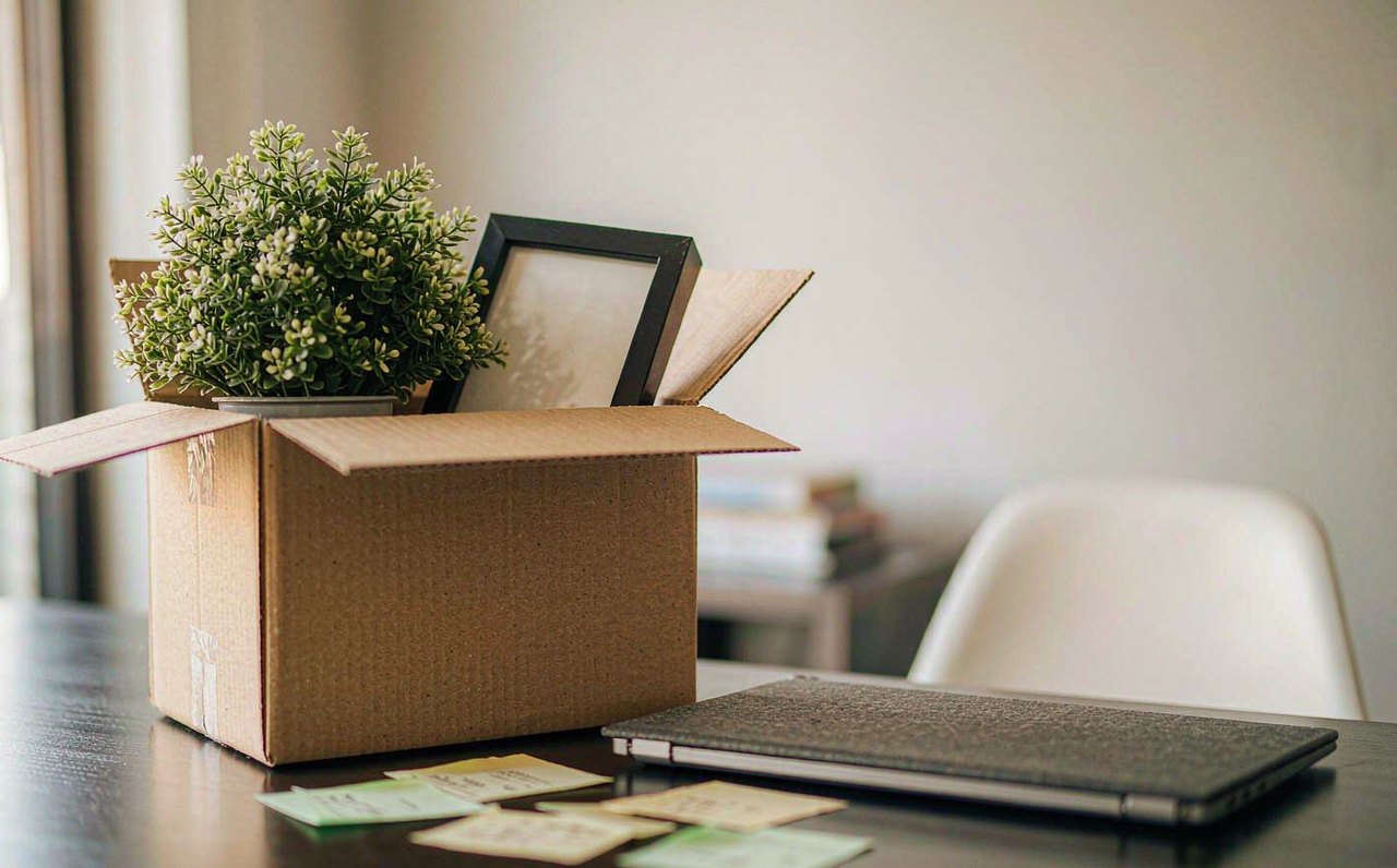 A cardboard moving box with a plant and picture frame on a desk beside a closed laptop in an office
