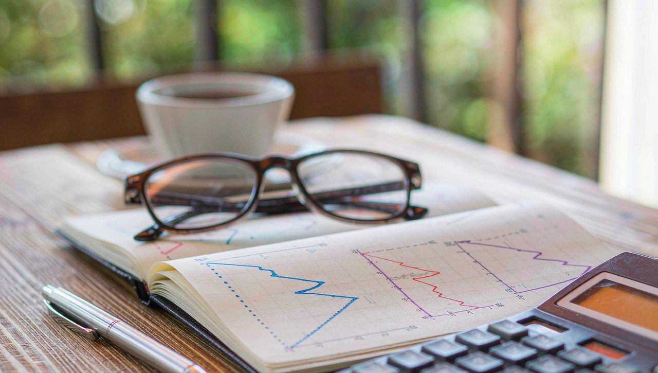 A calculator, a pen, and a notebook with hand-drawn simple charts on a wooden desk in soft daylight, a cup of tea nearby
