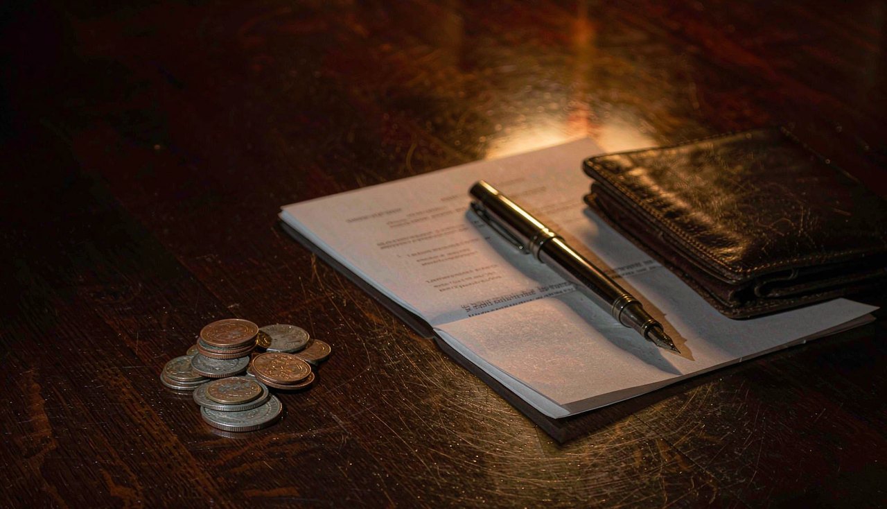 Coins arranged in neat columns beside a printed share certificate and a fountain pen on a dark wooden table